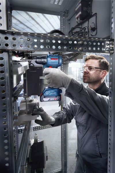 A person wearing safety equipment uses a cordless drill inside an electrical cabinet.