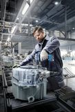 A person wearing safety equipment uses a cordless drill to assemble machinery in a factory.