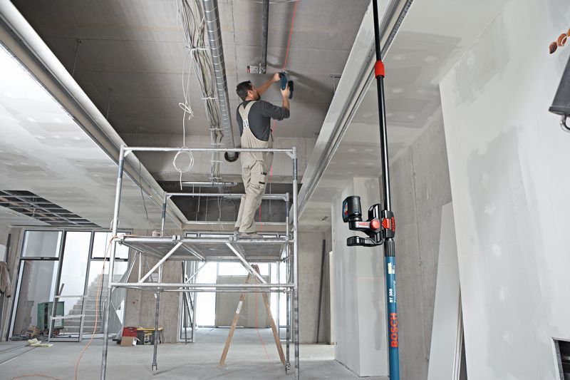 A person stands on scaffolding installing ductwork, guided by a laser leveling tool on a universal mount.