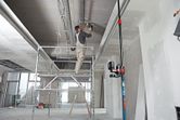 A person stands on scaffolding installing ductwork, guided by a laser leveling tool on a universal mount.