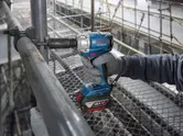 A person wearing safety equipment uses a cordless impact wrench on a metal pipe.