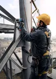 A person wearing safety equipment uses a cordless drill on a metal structure.