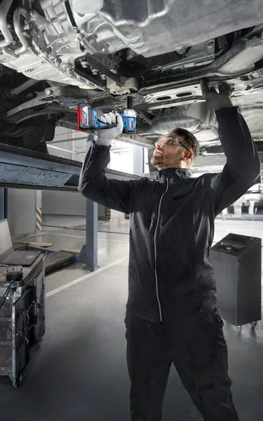 A person wearing safety equipment uses a power drill to work underneath a raised vehicle.