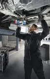 A person wearing safety equipment uses a cordless impact wrench to tighten bolts under a car.