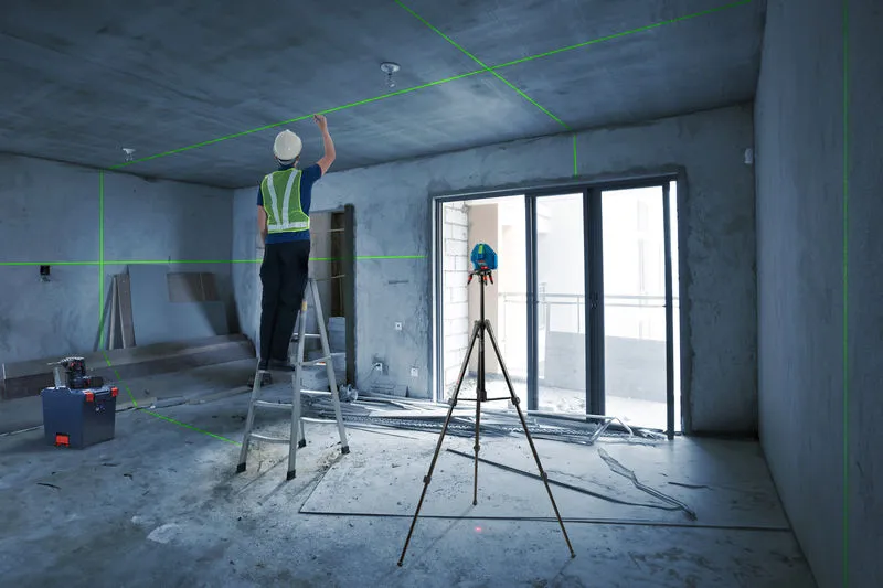 A person wearing safety equipment adjusts the ceiling using a laser leveling tool on a tripod.