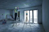 A person wearing safety equipment adjusts the ceiling using a laser leveling tool on a tripod.