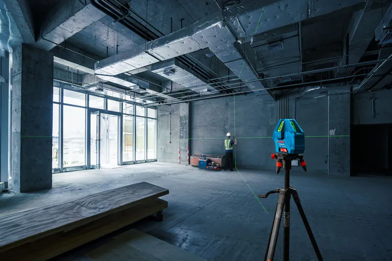 A laser leveling tool on a tripod projects green lines while a person wearing safety equipment marks a wall.