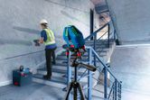 A person wearing safety equipment uses a power drill while a laser leveling tool projects a green line.