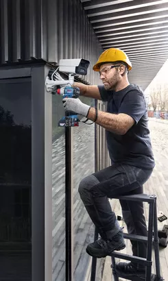 Professional Promotion Set of Combo GDX 18V-285 + GSB 185-LI with 2x2.0Ah, GAL 18V-20 in Case A person wearing safety equipment installs a security camera using a power drill.