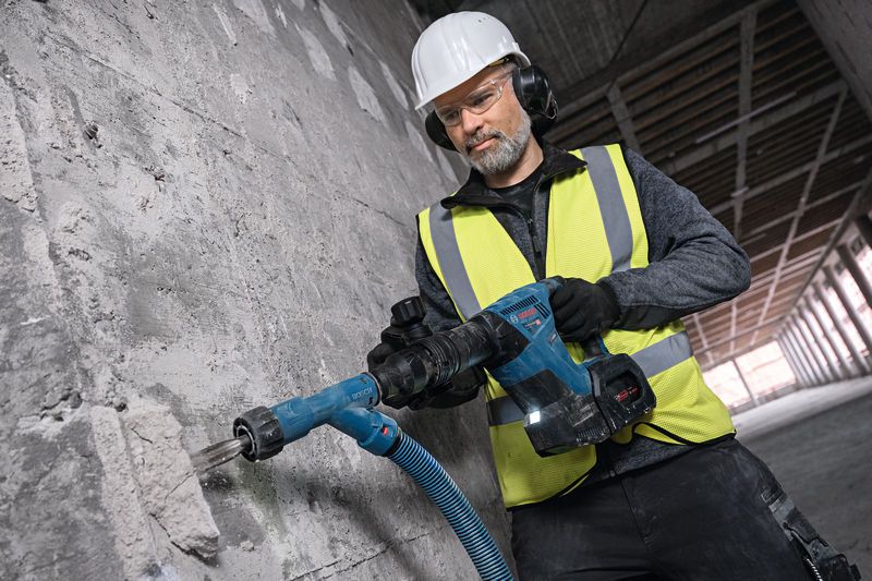 A worker wearing safety equipment uses a cordless demolition hammer on a concrete wall.