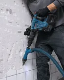 A person wearing safety equipment uses a cordless demolition hammer to break wall tiles.