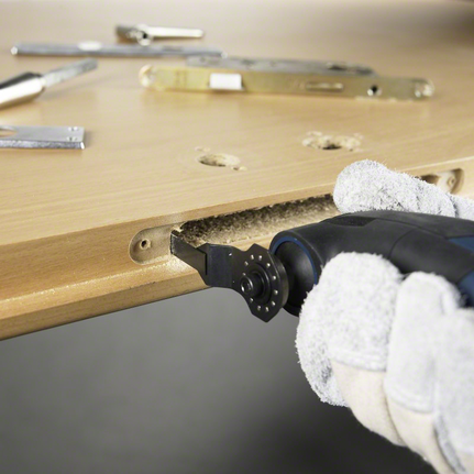 Person wearing safety equipment uses a multi-tool to cut a rectangular slot in a wooden door.