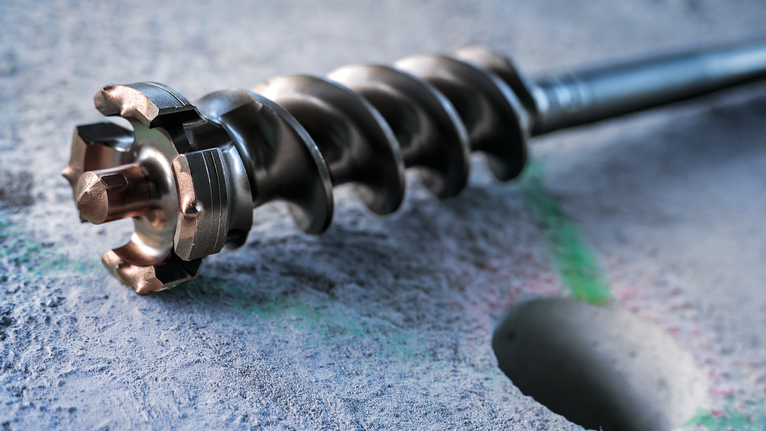 Large drill bit resting on concrete near a freshly drilled hole.