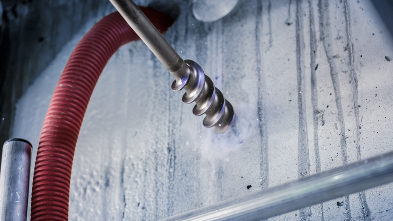 A rotary hammer drill bit makes a hole in a concrete wall with a vacuum hose attached.