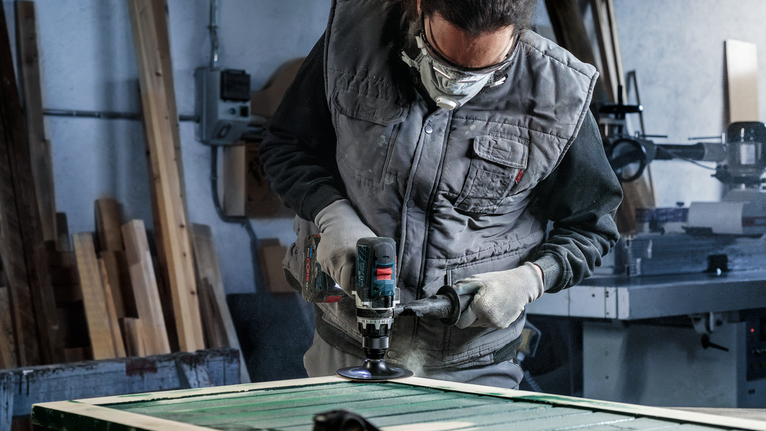 Person wearing safety equipment polishes a wooden surface with a power tool.