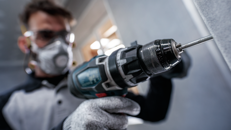 Person wearing safety equipment drills into a wall with a power tool.