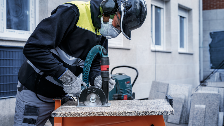 Person wearing safety equipment cuts a stone slab with a circular power saw.