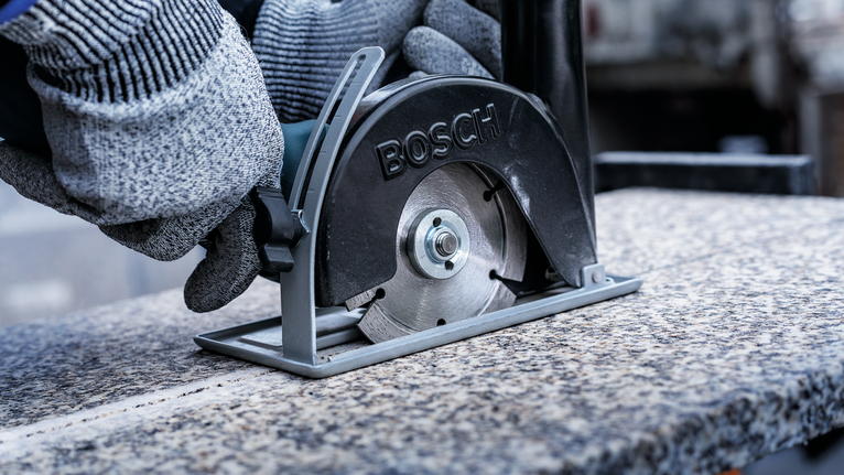 A person wearing safety equipment uses a metal saw to cut a granite slab.