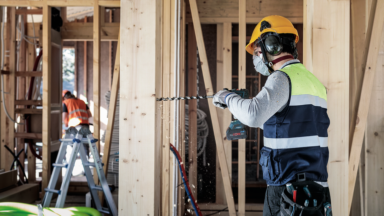 A person wearing safety equipment drills into a wooden frame at a construction site.