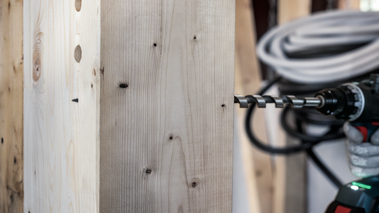 Person wearing safety equipment drills through thick wooden plank with a power tool.