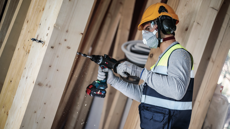 Person wearing safety equipment drills into a wooden beam.