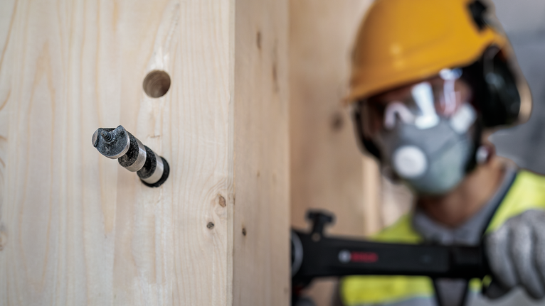 A person wearing safety equipment drills into a wooden beam.