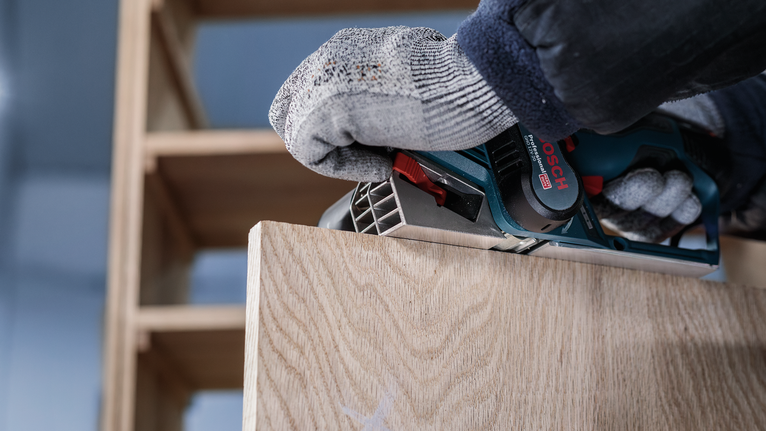 Person wearing safety equipment uses a power planer on a wooden board.