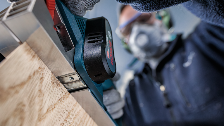 Person wearing safety equipment uses a power planer on a wooden board.