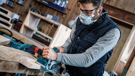Person wearing safety equipment uses a power planer to smooth wooden boards.