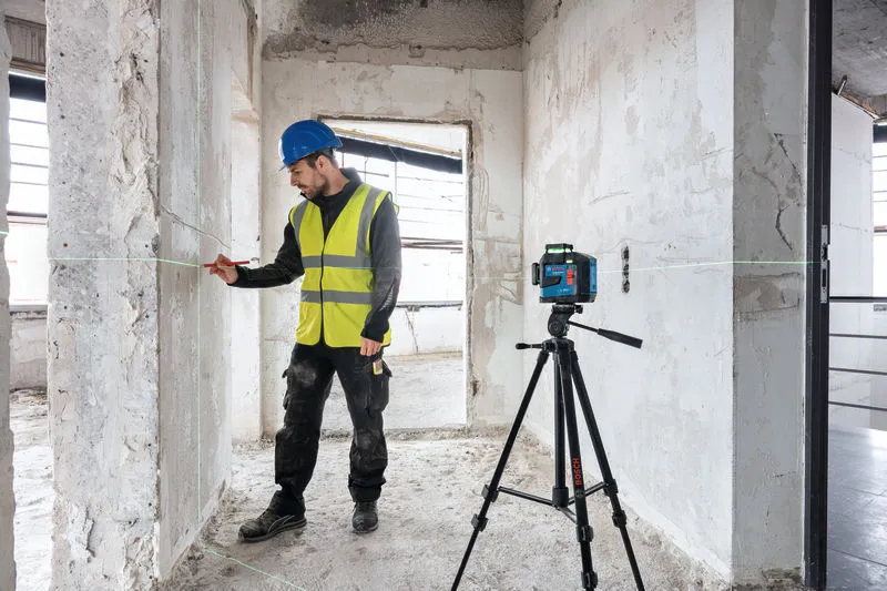 A person wearing safety equipment marks a wall guided by a laser leveling tool on a building tripod.