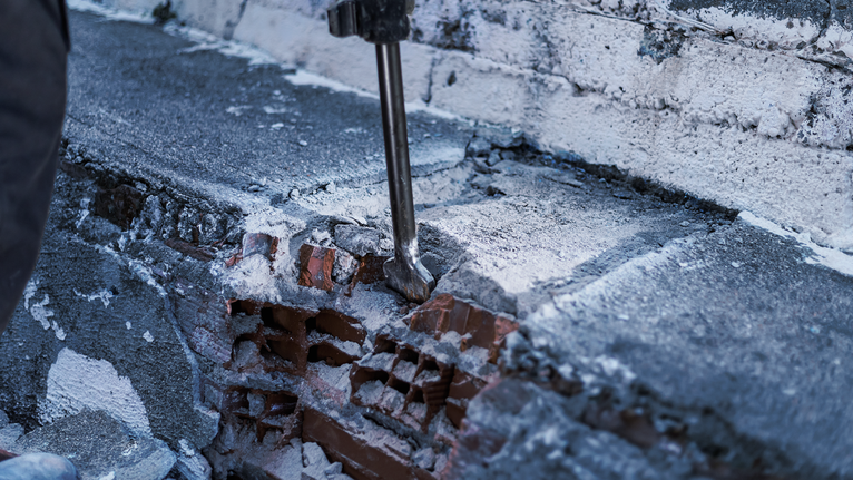 Demolition tool breaking concrete on a brick wall.