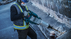 A person wearing safety equipment uses a demolition hammer to break concrete.