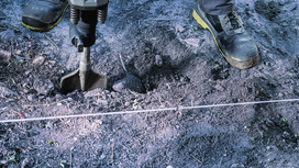 Worker wearing safety equipment uses a demolition hammer to break soil.