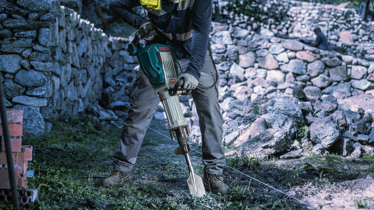 Person wearing safety equipment uses a demolition hammer to break ground near a stone wall.