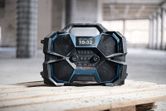 Portable construction site radio with digital display sits on wooden planks in a building under construction.
