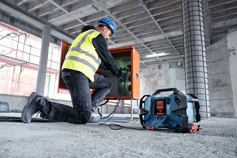 A person wearing safety equipment connects wires at a construction site near a jobsite radio.