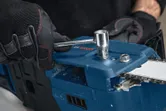 A person wearing safety equipment adjusts a blue chainsaw with a socket wrench.