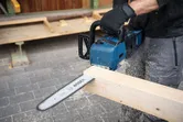 A person wearing safety equipment cuts a wooden beam with a blue chainsaw.