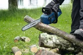 A person wearing safety equipment cuts logs outdoors using a cordless chainsaw.