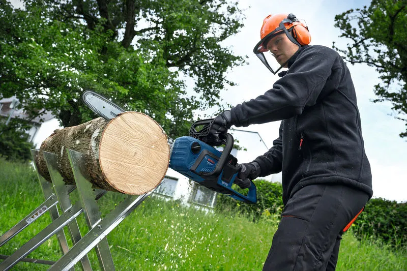 A person wearing safety equipment uses a cordless chainsaw to cut a log outdoors.