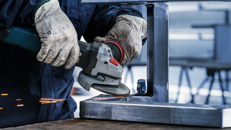 A person wearing safety equipment grinds a metal beam with an angle grinder.