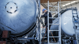 A person wearing safety equipment uses a power tool on a large metal tank.