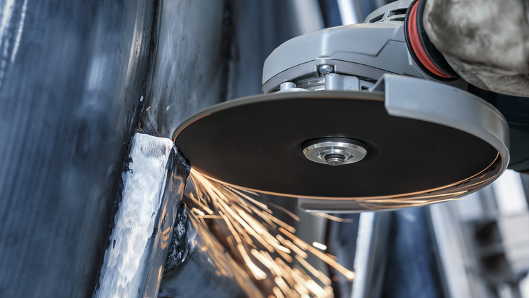 Person wearing safety equipment uses an angle grinder to cut metal, sparking.