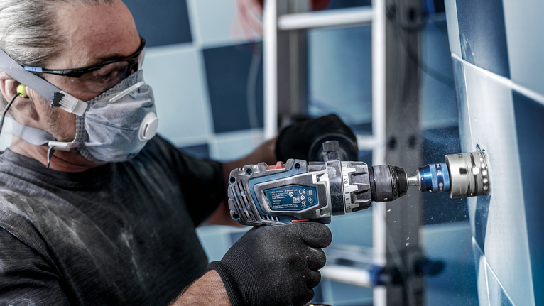 A person wearing safety equipment drills a circular hole in a tiled wall.