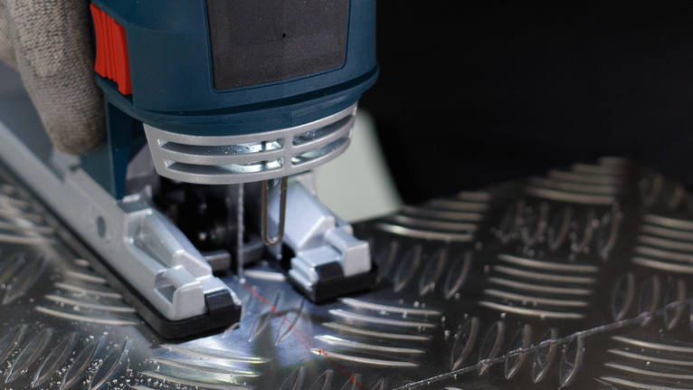 PRO Alu T127D Jigsaw Blade, 100 mm, 5 pcs Person wearing safety equipment operates a jigsaw to cut a patterned metal sheet.
