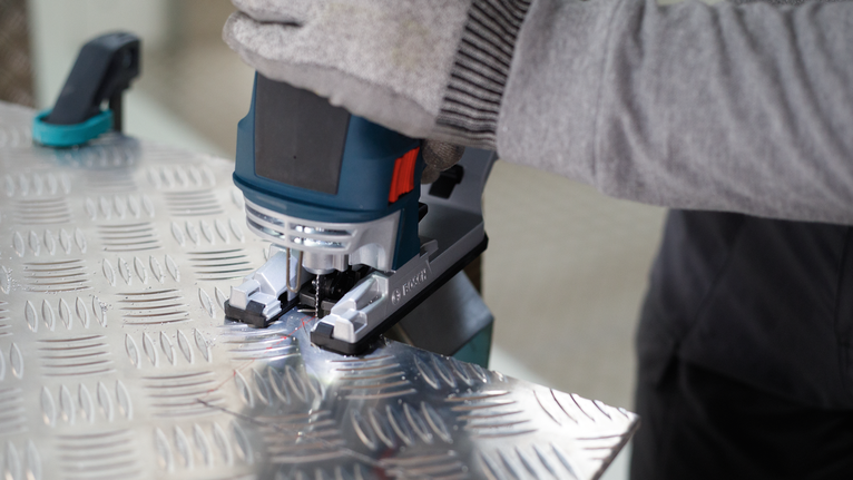 PRO Alu T127D Jigsaw Blade, 100 mm, 5 pcs Person wearing safety equipment uses a jigsaw to cut patterned metal sheet.