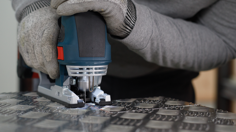 PRO Alu T127D Jigsaw Blade, 100 mm, 5 pcs Person wearing safety equipment guides a jigsaw to cut patterned metal sheet.