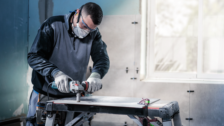 Person wearing safety equipment cutting tile with a power tool on a workbench.