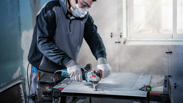 Person wearing safety equipment uses a power tool to cut a large tile sheet.
