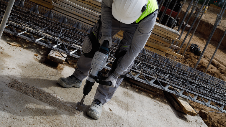 A person wearing safety equipment drills into concrete at a construction site.